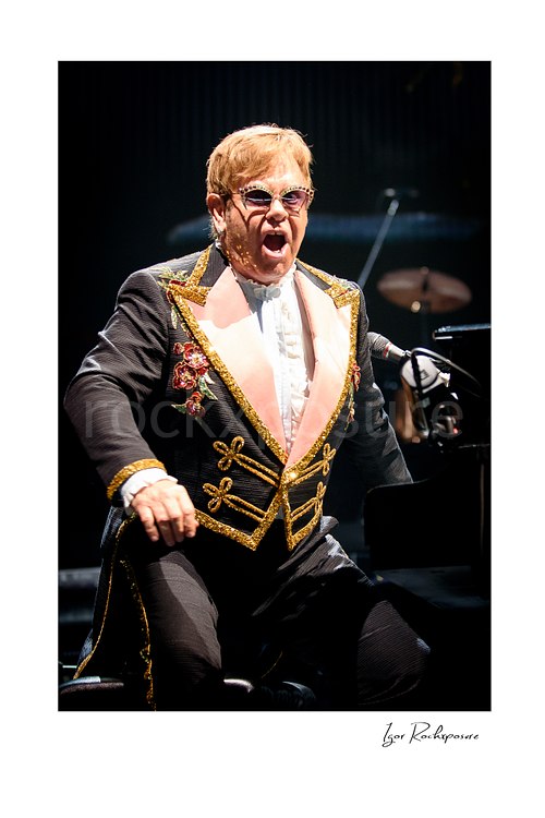 Vertical color image of Elton John seated at the piano in a jeweled stage suit with an intense singing expression