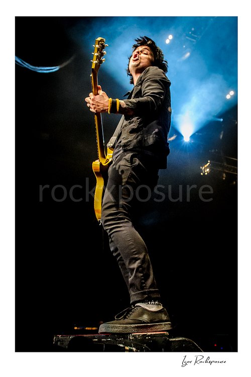 Vertical color image of Billie Joe Armstrong of Green Day playing guitar on a stage riser under blue concert light