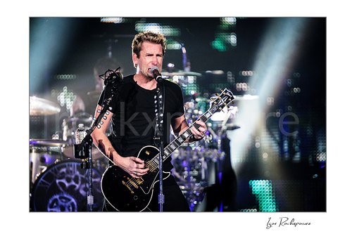 Horizontal color image of Chad Kroeger of Nickelback singing into a microphone while playing guitar with the drummer visible behind him under bright concert beams