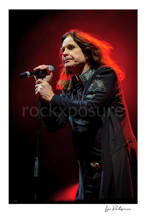 Vertical concert photography of Ozzy Osbourne of Black Sabbath singing into a microphone under dramatic red stage lighting