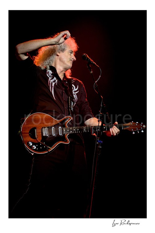 Vertical color image of Brian May of Queen holding his guitar beside a microphone under warm red concert light