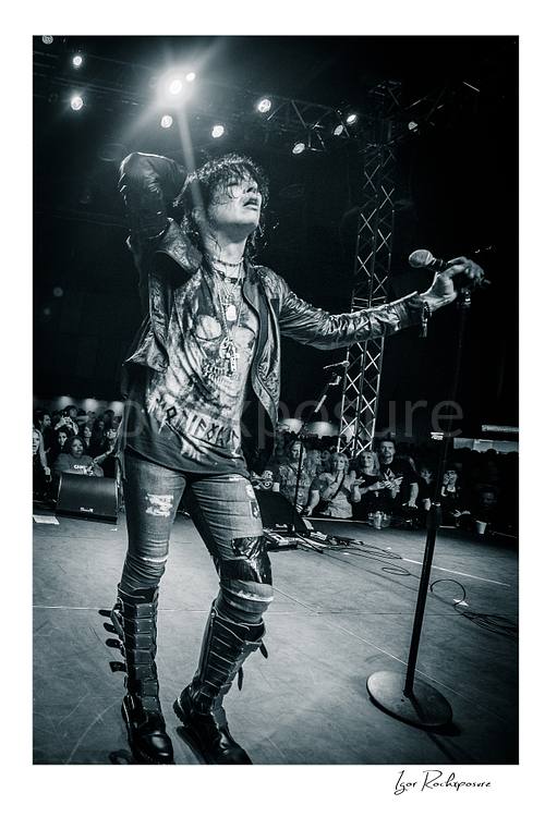 Vertical black and white image of Tom Keifer performing live on stage with one hand behind his head and the other on a microphone stand, with the crowd visible behind him