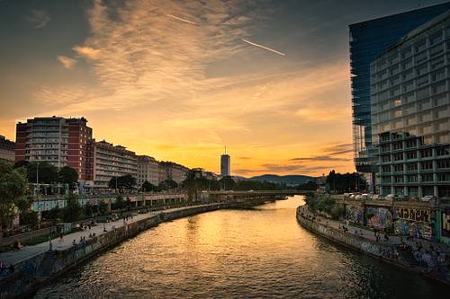 Weitwinkelaufnahme des Wiener Donaukanals bei einem goldenen Sonnenuntergang im Juni. Der orange leuchtende Himmel spiegelt sich im Wasser, flankiert von graffiti-verzierten Uferpromenaden, einem roten Wohngebäude links und modernen Glasfassaden rechts. Blick von der Marienbrücke.