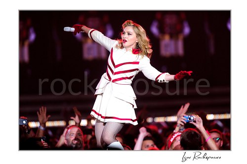 Horizontal color image of Madonna performing in a white and red stage outfit while moving through the crowd with a microphone in hand