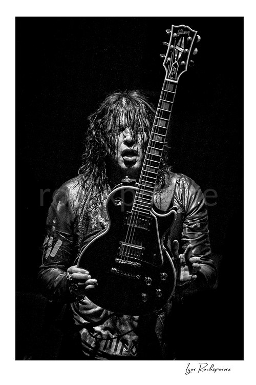 Vertical black and white image of Tom Keifer performing live on stage, holding a black guitar upright in front of him against a dark background