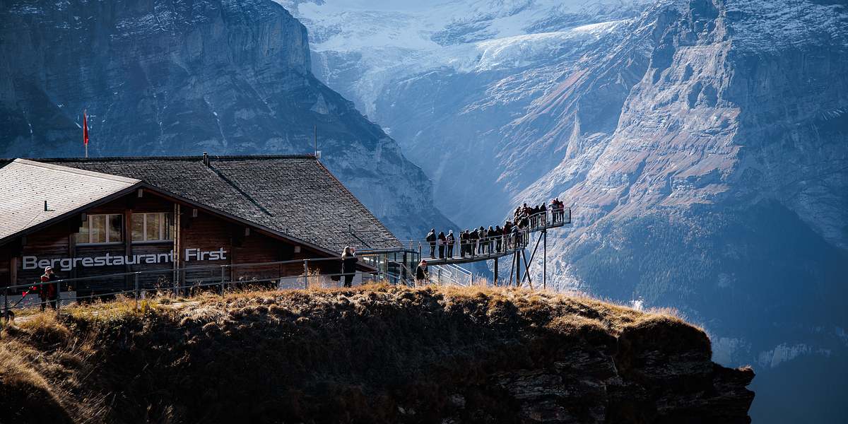 Bergrestaurant First auf dem Grat mit Besuchern auf dem auskragenden Cliff Walk, dahinter schneebedeckte Gipfel und Gletscher im Nachmittagslicht