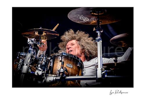 Horizontal color image of Tommy Aldridge of Whitesnake performing live on stage behind the drum kit, captured in a close-up view between cymbals and drums