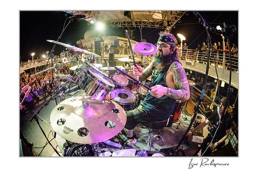 Horizontal color image of Mike Portnoy playing drums on stage at night in a wide fisheye concert scene with the crowd and ship deck behind him