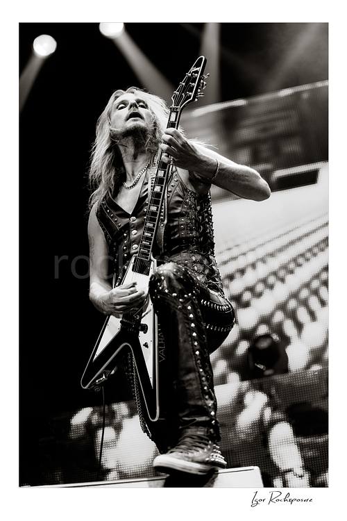 Vertical black and white image of Richie Faulkner of Judas Priest performing live on stage, playing flying V guitar in a dramatic low-angle shot under bright concert lighting