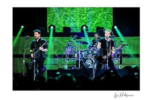 Horizontal color image of Ryan Peake and Chad Kroeger of Nickelback performing onstage with Daniel Adair visible behind them under bright green concert lighting