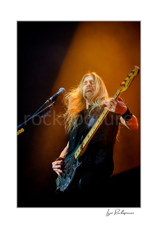 Vertical color image of James LoMenzo of Megadeth playing bass on stage with long hair and warm spotlight