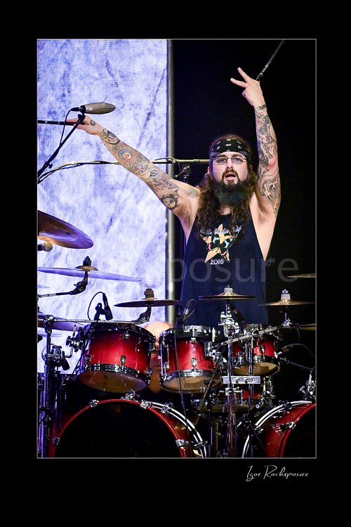 Vertical color image of Mike Portnoy at the drum kit with both tattooed arms raised and a drumstick in hand under bright stage lighting
