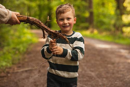 Ein kleiner Junge läuft lächelnd auf einem Waldweg und hält stolz ein großes, verzweigtes Stück Holz in den Händen.