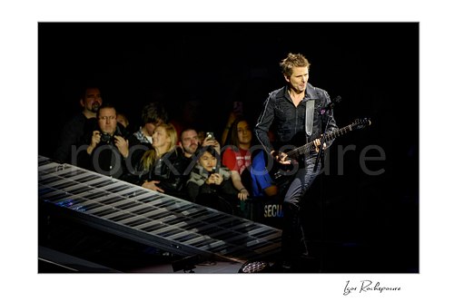 Horizontal color image of Matt Bellamy of Muse playing guitar beside a piano-key stage ramp before the arena crowd