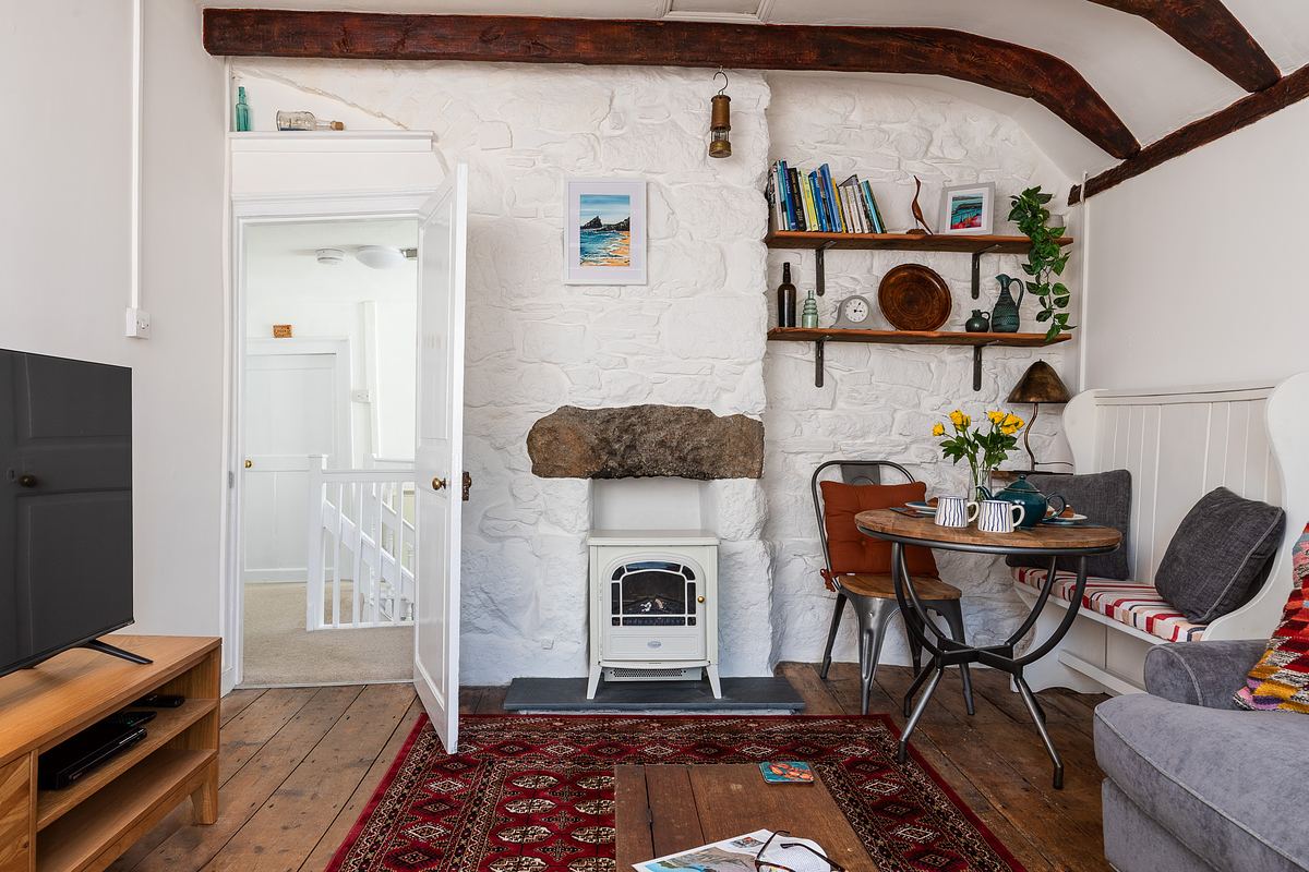 A cozy living area with white stone walls, exposed wood beams, a small dining table, shelves with books and decor, and a white stove heater on a red rug.