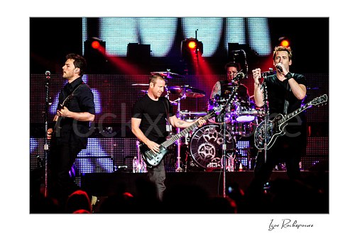Horizontal color image of Nickelback performing onstage with Ryan Peake, Mike Kroeger, Chad Kroeger, and Daniel Adair under red concert lighting