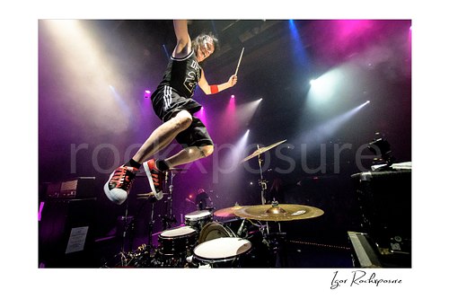 Horizontal color image of Zoltan Chaney leaping above his drum kit under purple and white stage lights
