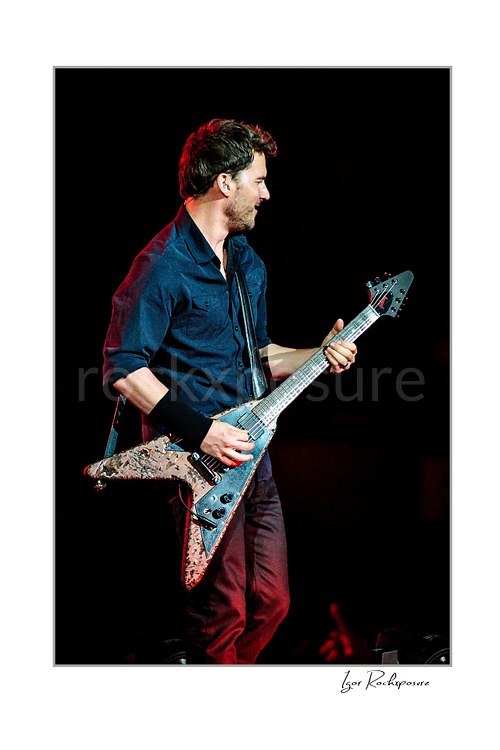 Vertical color image of Ryan Peake of Nickelback playing guitar onstage under red concert light