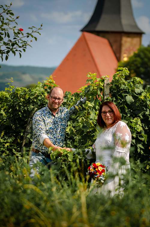 Brautpaar steht vor grünen Sträuchern unterhalb der Burg Weibertreu, im Hintergrund ein Kirchturm mit rotem Dach