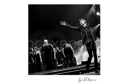 Square black and white image of Mick Jagger of The Rolling Stones performing beside a choir on stage during You Can’t Always Get What You Want