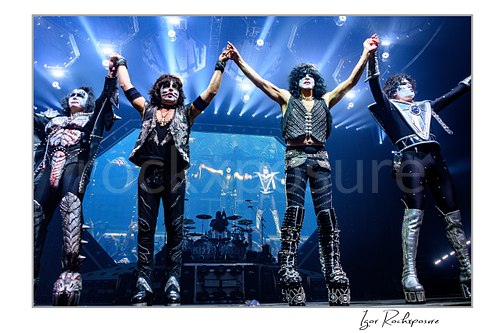 Horizontal color photograph of the full KISS band in full makeup and costumes standing with their backs to the stage and their hands raised toward the crowd as bright concert lighting fills the finale moment