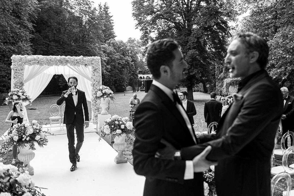Sebastien CLAVEL Photographe mariage Lyon e mari&eacute; en costume boit une coupe de champagne en marchant seul sous l'arche de mariage, tandis que les invit&eacute;s et les musiciens se tiennent en arri&egrave;re-plan