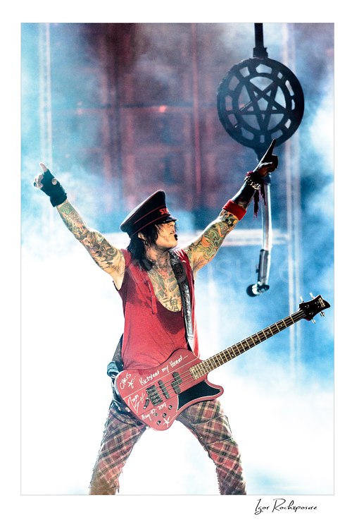 Vertical color image of Nikki Sixx performing with bass guitar during a Mötley Crüe concert, raising both arms in blue stage smoke