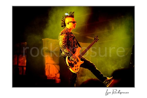 Horizontal color image of Johnny Christ of Avenged Sevenfold playing bass onstage in sunglasses with a spiked mohawk and glowing yellow stage haze