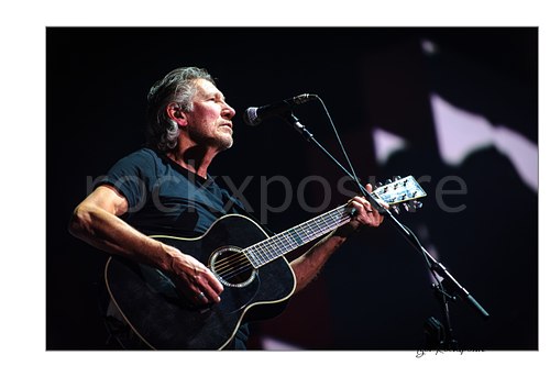 Roger Waters stands at a microphone strumming a black acoustic guitar, shown in a horizontal concert photograph with pale shapes in the dark background.