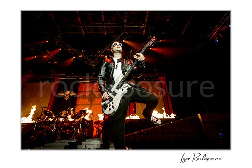 Horizontal color image of Johnny Christ of Avenged Sevenfold playing bass onstage with flames, red lighting, and the drummer visible behind him.