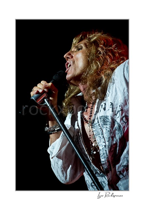 Vertical color close-up image of David Coverdale of Whitesnake performing live on stage, singing into a microphone while holding a mic stand against a dark background