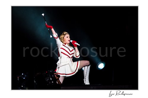 Horizontal color image of Madonna singing into a microphone in a white and red stage outfit while raising a baton under dramatic spotlight