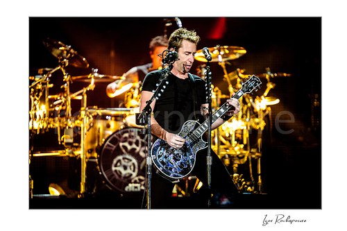 Horizontal color image of Chad Kroeger of Nickelback playing guitar onstage with the drummer visible behind him under warm concert lighting