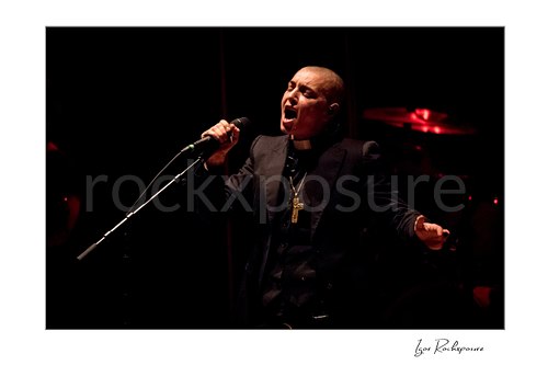Sinéad O’Connor sings into a microphone with eyes closed and her free hand extended, wearing a black jacket and cross necklace before red-lit cymbals.