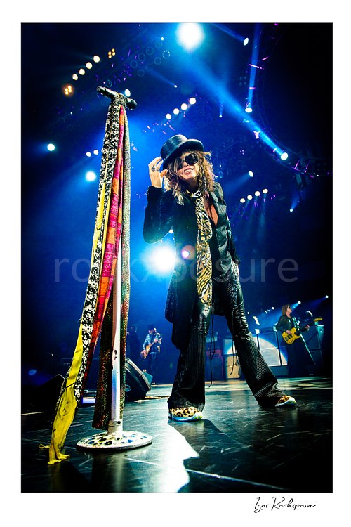 Vertical color image of Steven Tyler of Aerosmith performing live on stage in a black top hat with a scarf-wrapped microphone stand, patterned scarf, and blue concert lighting
