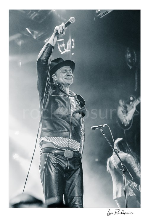 Vertical black and white image of Gord Downie of The Tragically Hip raising a microphone high above his head, wearing a hat and leather jacket in bright concert haze with a blurred band member behind him