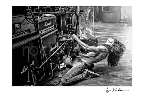 Horizontal black and white image of Brent Woods playing guitar shirtless on the stage floor beside Marshall amplifier stacks