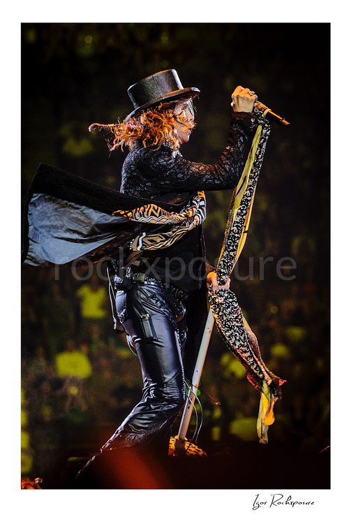 Vertical color image of Steven Tyler of Aerosmith performing live on stage in a black top hat with a scarf-wrapped microphone stand, flowing hair, and black leather pants