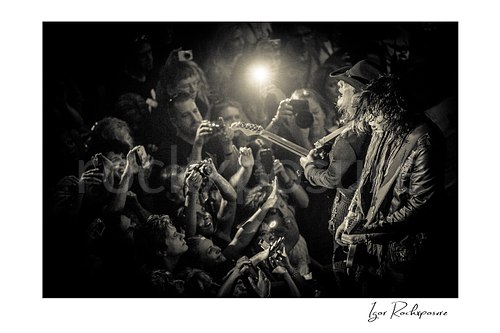 Horizontal black and white image of Tom Keifer and Jeff LaBar performing live at the edge of the stage, surrounded by fans reaching up with cameras and bright flash light behind them