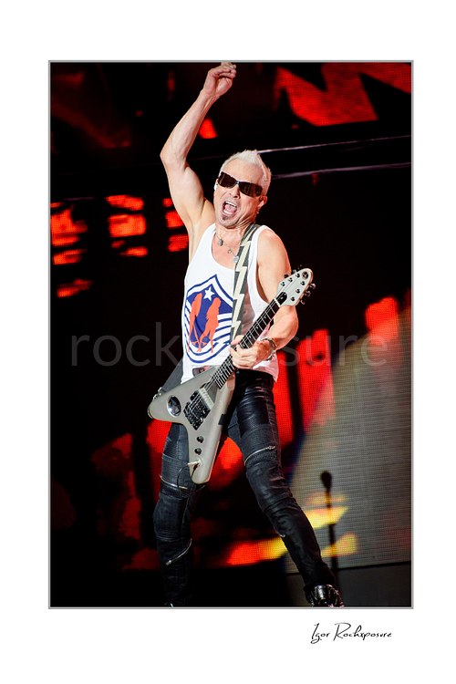 Vertical color image of Rudolf Schenker of Scorpions performing live with silver guitar, raised arm, and vivid red stage lights