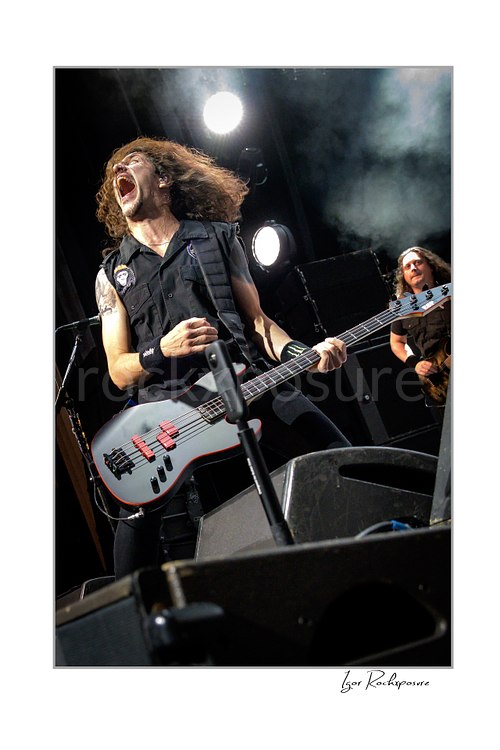 Vertical color image of Frank Bello of Anthrax performing live on stage, shouting while playing bass guitar with another band member visible in the background under bright concert lights