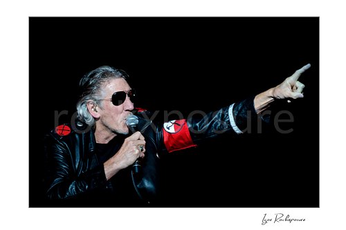 Roger Waters points diagonally upward while holding a microphone to his mouth, wearing sunglasses, a black leather jacket, and a red armband.