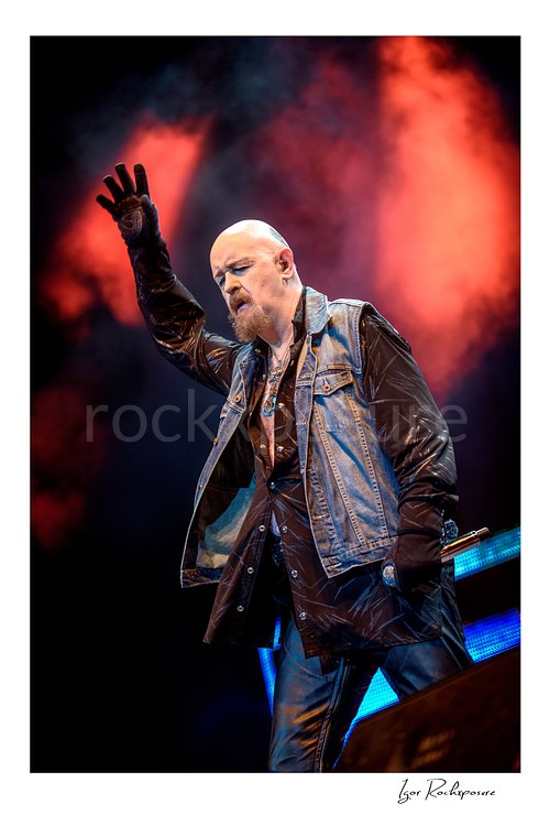Vertical color image of Rob Halford of Judas Priest performing live on stage with one gloved hand raised under dramatic red smoke and blue stage lighting