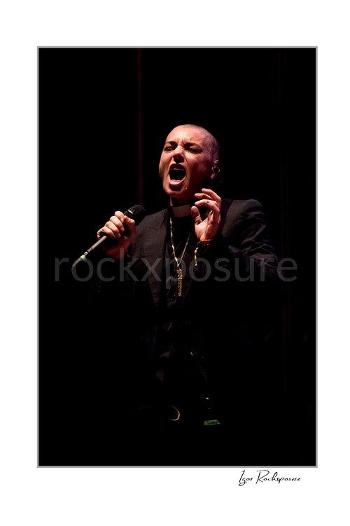 Sinéad O’Connor sings with eyes closed and mouth open, holding a microphone and raising her free hand near her face against a dark stage background.