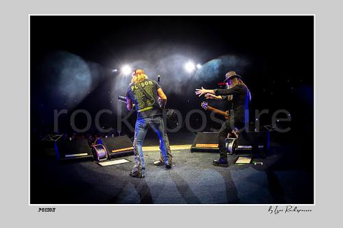 Horizontal color image of Bret Michaels and C.C. DeVille on a Poison stage, photographed with a remote camera mounted on the drum kit facing forward, with bright backlights and stage smoke
