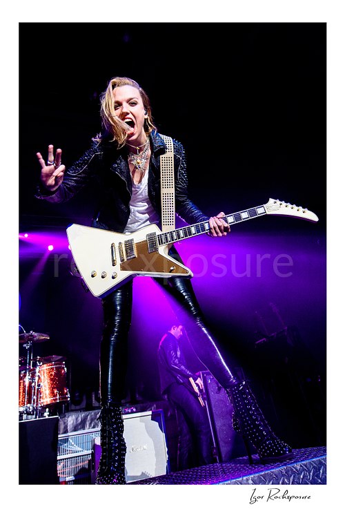 Vertical color image of Lzzy Hale of Halestorm performing live with a white guitar, flashing a peace sign and singing under vivid purple stage light