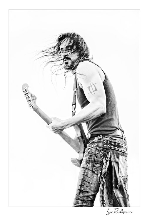 Vertical black and white image of Nuno Bettencourt performing live on stage with an electric guitar and long hair blowing across his face