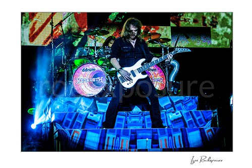 Horizontal color image of David Ellefson of Megadeth playing bass on stage with the drummer visible behind him and bright blue concert lighting