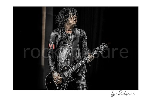 Horizontal color image of Tom Keifer performing live on stage with a black guitar, wet hair falling across his face against a dark concert background