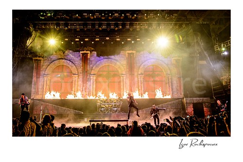 Horizontal color image of Avenged Sevenfold performing on stage in a dramatic full-band wide shot with fire, stage smoke, and a large crowd
